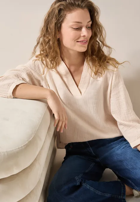 Frau mit lockigem Haar sitzt entspannt auf dem Sofa, trägt helle Bluse und Jeans.